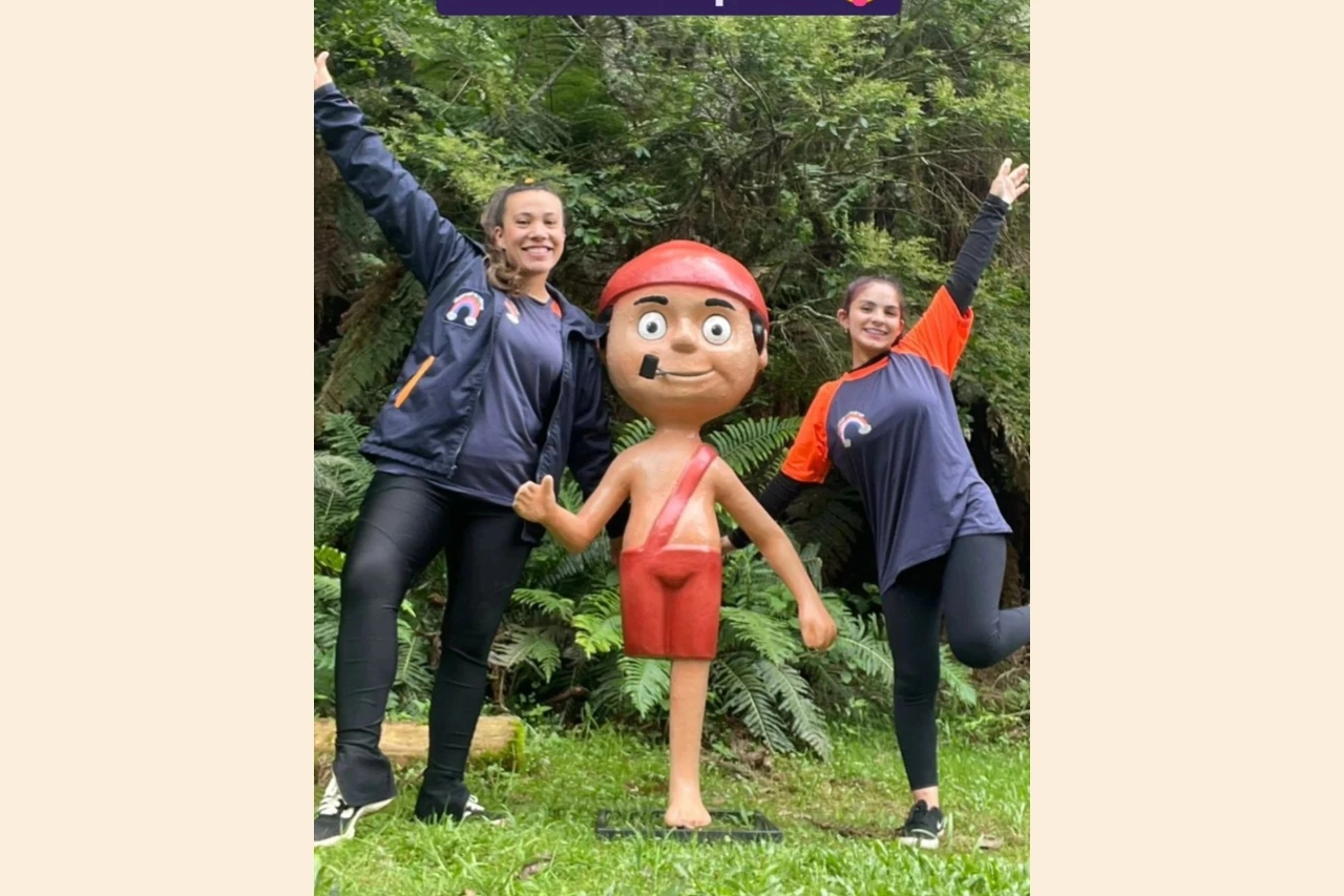 Duas pessoas ao lado de uma figura do Saci em área arborizada.