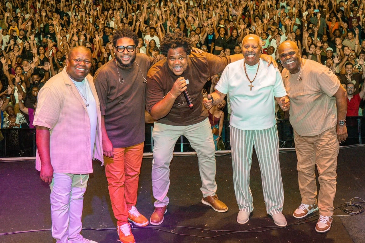 Cinco integrantes do Fundo de Quintal no palco, com o público ao fundo.
