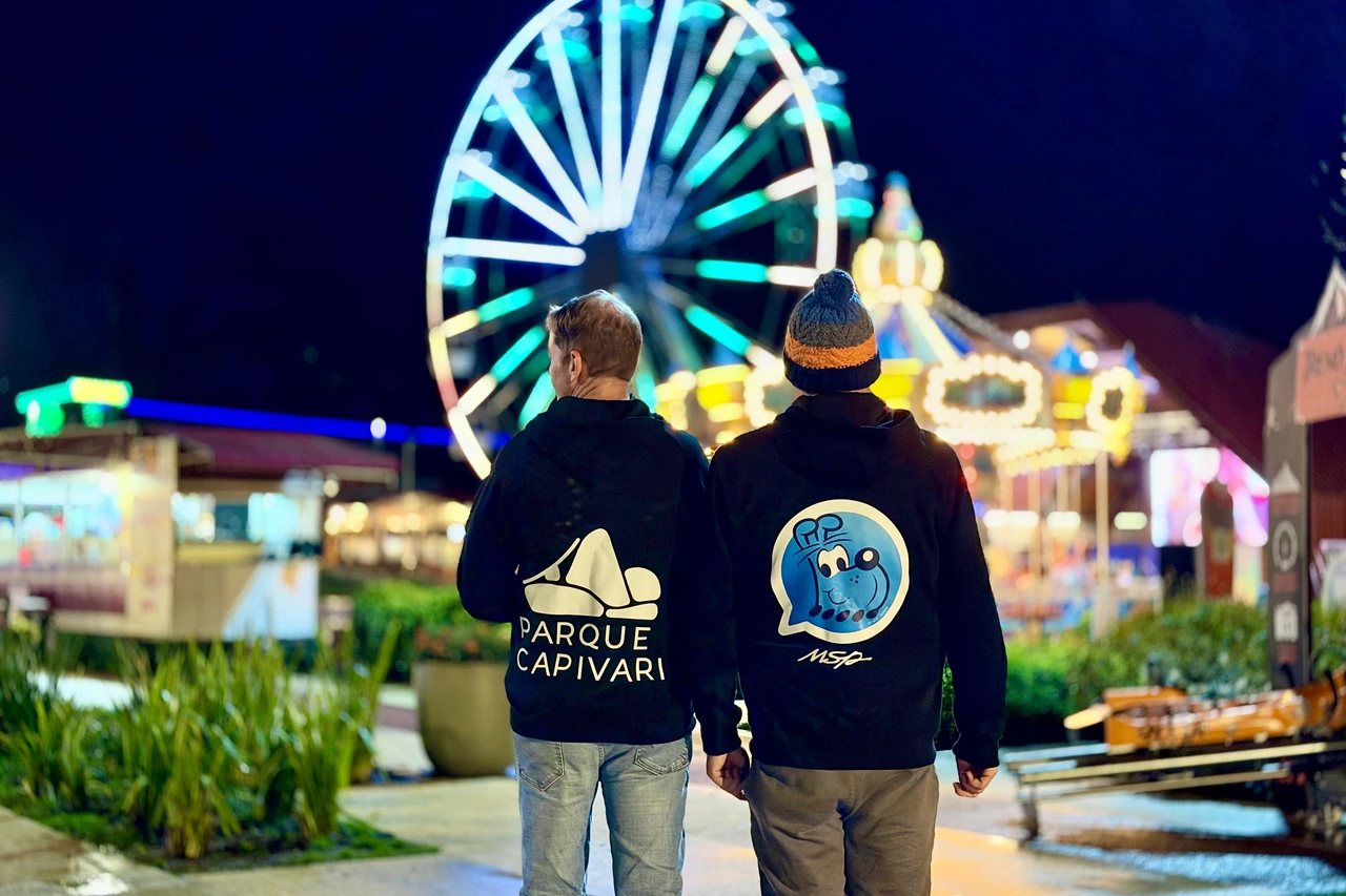 Duas pessoas de costas, com marcas do Parque Capivari e da MSP nas roupas, diante da roda-gigante iluminada.